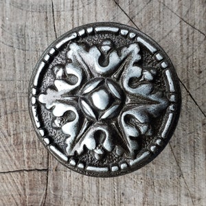 May include: A silver metal knob with a decorative floral design. The knob has a circular shape and a raised rim.