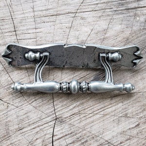 Puede incluir: Un tirador de cajón de metal plateado con un diseño decorativo y ornamentado. El tirador tiene una placa rectangular con la parte superior e inferior curvadas, y dos asas que se extienden desde la placa. Las asas también están decoradas con detalles ornamentados.