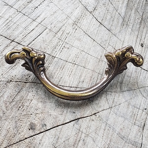 May include: A vintage-style brass drawer pull with ornate floral detailing. The curved handle is attached to decorative end pieces with a scroll design. The hardware has a warm, aged finish, suggesting a classic or antique aesthetic. The pull is designed for furniture.