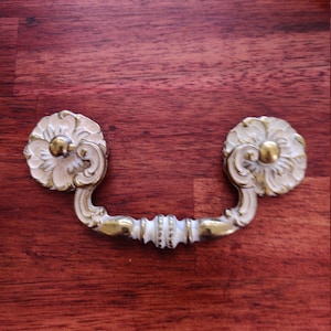 May include: An ornate white and gold drawer pull with a decorative floral design. The pull is attached to a wooden surface.