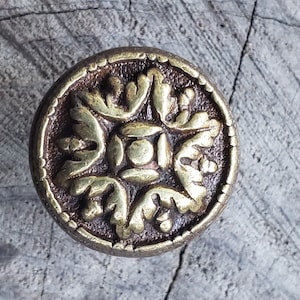 May include: A round, brass-colored button with a decorative, floral design. The button has a raised, central square with a geometric pattern.