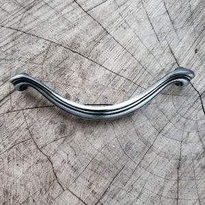 May include: A silver cabinet pull with a curved, arched design. The handle has decorative, scrolled ends and three parallel grooves. The hardware is set against a weathered, wooden background.