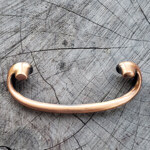 May include: A copper-colored cabinet pull with a curved handle and rounded ends. The hardware is designed for easy gripping and is likely used for drawers or cabinet doors. The pull is made of metal and has a smooth, reflective surface.