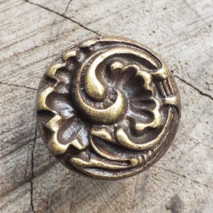 May include: A round, ornate brass knob with a swirling, floral design. The knob has a dark brown patina and a gold-toned accent.
