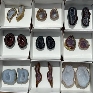 May include: Assortment of polished agate geodes in white cardboard boxes. The geodes display a variety of colors, including blue, purple, brown, and black, with crystalline interiors. Each geode is unique in shape and size.