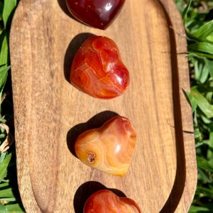 May include: Four heart-shaped stones in shades of red, orange, and yellow are arranged on a wooden tray. The stones have a polished, glossy finish, and the tray is oval-shaped. The background features green grass.