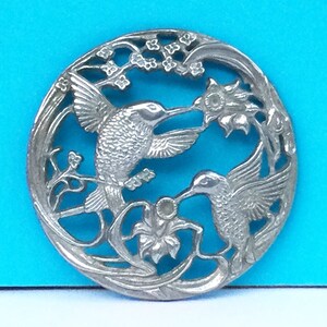 Könnte beinhalten: Silberner Anhänger mit zwei Kolibris im Flug, mit detaillierten Flügeln und Schnäbeln, umgeben von zarten floralen Designs.