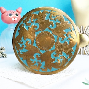 Op de afbeelding: Een ronde, vintage compactpoederdoos met een goudkleurig metalen frame en een turquoise centrum. Het centrum heeft een sierlijk, reliëf ontwerp in goud, met een centraal medaillon en wervelende bladerpatronen. Een klassieker.