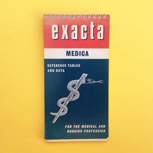 Op de afbeelding: Een blauw en rood spiraalgebonden notitieboek met de tekst "exacta medica" en "reference tables and data" gedrukt op de voorzijde. De omslag heeft ook een witte illustratie van een slang die om een stok is gewikkeld, een symbool van de geneeskunde.