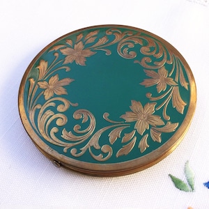 Op de afbeelding: Ronde, vintage compact met een teal emaille afwerking en gouden bloemen filigraan ontwerp. De compact heeft een goudkleurig metalen frame en een scharnierende opening. Een decoratief item.