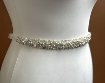 May include: A white beaded bridal belt with a satin ribbon. The belt is adorned with pearls and rhinestones.