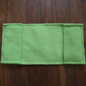 May include: A rectangular, light green fleece pad with stitched edges. The pad has two vertical seams dividing it into three sections. The item is displayed on a dark brown wooden surface.