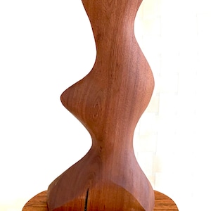 May include: A handcrafted wooden sculpture with a flowing, organic form. The sculpture is made of dark brown wood and is mounted on a lighter brown wooden base. The abstract design features curves and angles.
