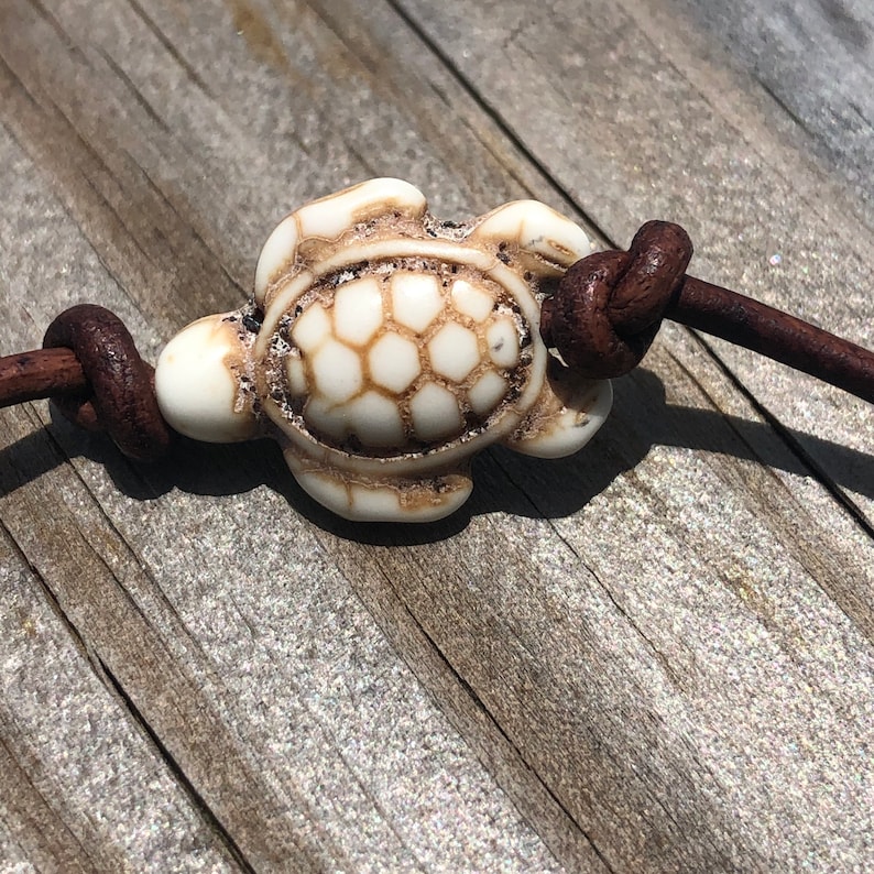 May include: A white ceramic turtle charm with a detailed shell design, attached to a brown leather cord.