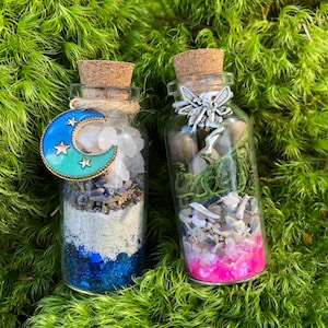 May include: Two small glass bottles filled with various items, including glitter, crystals, and a small fairy figurine. The bottle on the left has a blue crescent moon charm and blue glitter. The bottle on the right has pink glitter and a silver fairy charm.