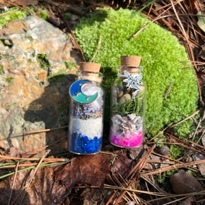 May include: Two small glass bottles filled with various items, including glitter, sand, and small shells. The bottle on the left has a blue crescent moon charm and the bottle on the right has a silver fairy charm.