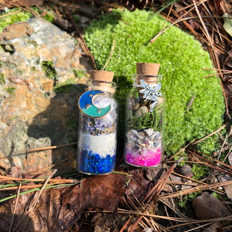 May include: Two small glass bottles filled with various items, including glitter, sand, and small shells. The bottle on the left has a blue crescent moon charm and the bottle on the right has a silver fairy charm.