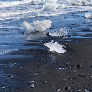 Puede incluir: Icebergs encallados en una playa de arena negra. Los icebergs son claros y transl&uacute;cidos, y la arena es oscura y h&uacute;meda. El oc&eacute;ano est&aacute; en el fondo, y las olas rompen en la orilla.