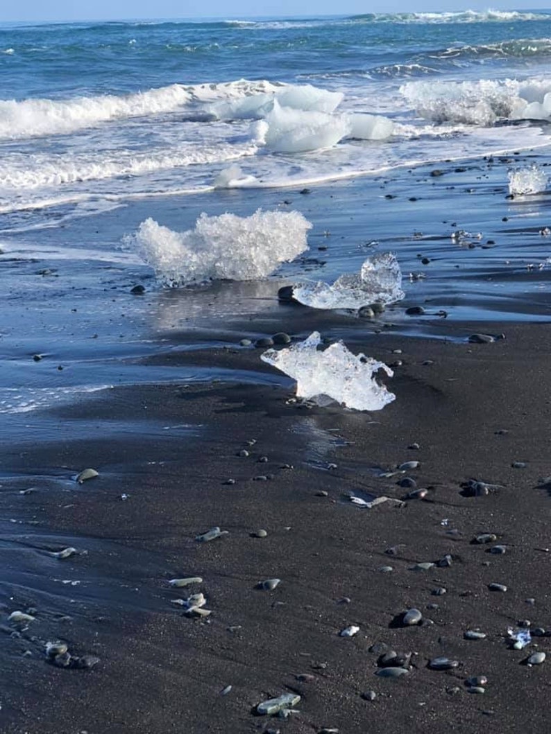 Puede incluir: Icebergs encallados en una playa de arena negra. Los icebergs son claros y transl&uacute;cidos, y la arena es oscura y h&uacute;meda. El oc&eacute;ano est&aacute; en el fondo, y las olas rompen en la orilla.