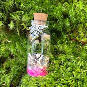 May include: A small glass bottle filled with various natural elements, including moss, small stones, and pink glitter. A silver fairy charm is attached to the cork stopper.