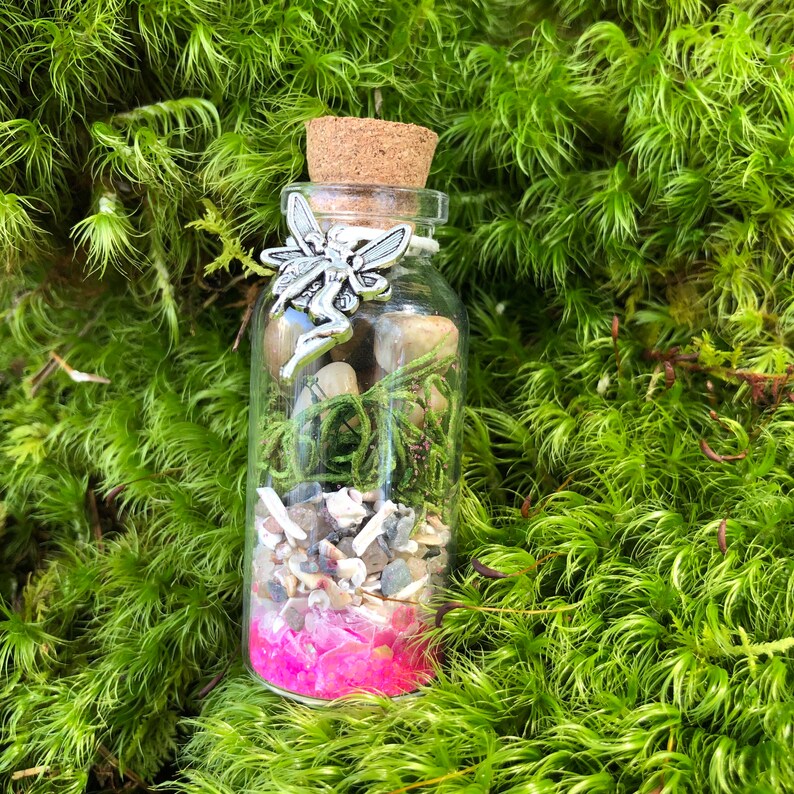 May include: A small glass bottle filled with various natural elements, including moss, small stones, and pink glitter. A silver fairy charm is attached to the cork stopper.