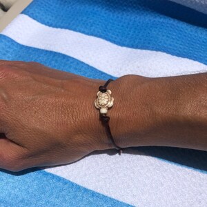 May include: A brown leather bracelet with a white turtle charm. The bracelet is tied with a knot and is worn on a person's wrist.
