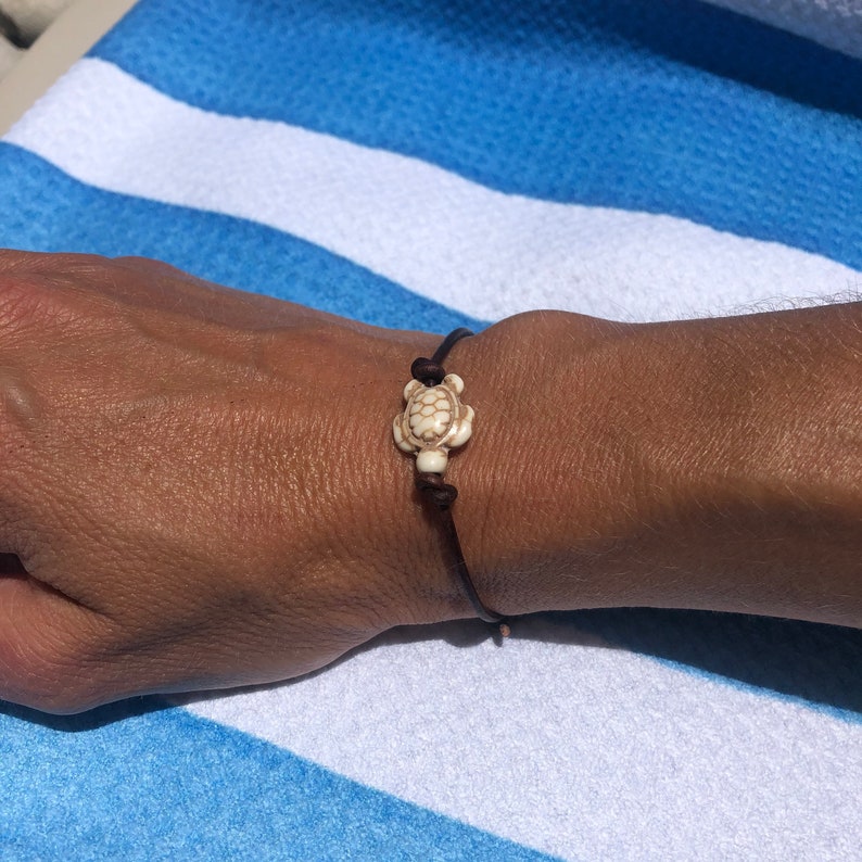 May include: A brown leather bracelet with a white turtle charm. The bracelet is tied with a knot and is worn on a person's wrist.