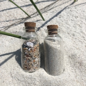 May include: Two small glass bottles with cork tops. One bottle is filled with white sand and the other is filled with small seashells.