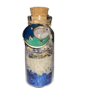 May include: A small glass bottle with a cork stopper filled with white glitter, blue glitter, dried herbs, and white crystals. A blue and green crescent moon charm with gold stars is attached to the bottle.