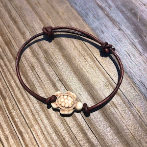 May include: A brown leather bracelet with a white turtle charm. The bracelet is tied with a knot on one end and has a small white bead on the other end.
