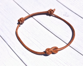 May include: A brown leather bracelet with a knotted design. The bracelet features a simple, adjustable design with knots at each end and in the center. The leather has a natural, worn appearance.