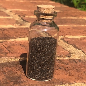 Puede incluir: Una peque&ntilde;a botella de vidrio con un tap&oacute;n de corcho llena de arena negra. La botella est&aacute; sobre una superficie de ladrillo.