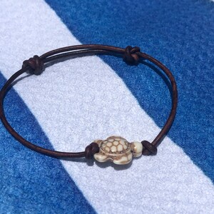 May include: A brown leather bracelet with a white turtle charm. The bracelet is tied with a knot and the turtle charm is in the center of the bracelet.