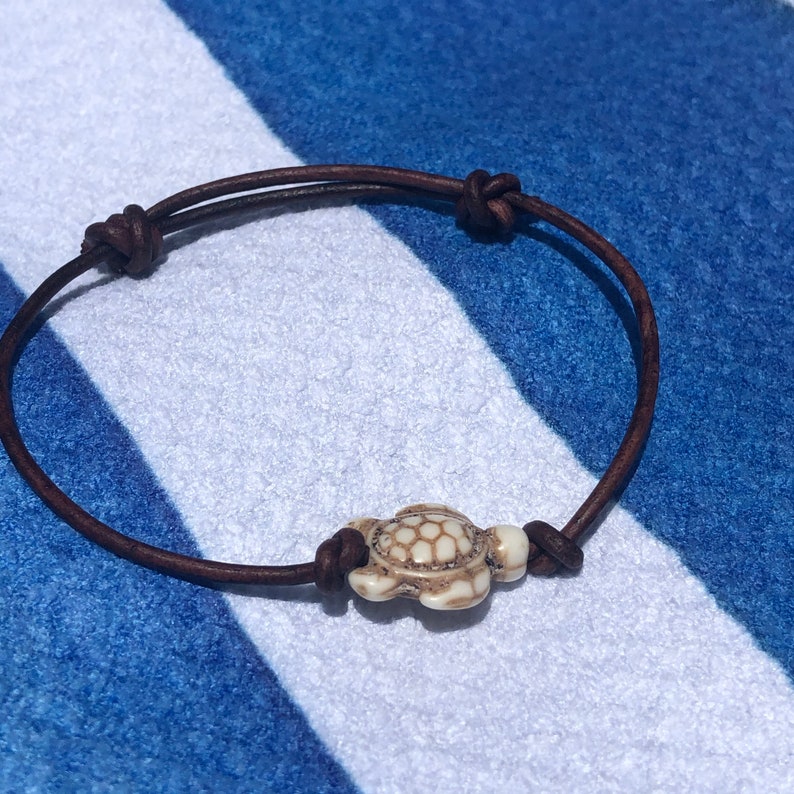 May include: A brown leather bracelet with a white turtle charm. The bracelet is tied with a knot and the turtle charm is in the center of the bracelet.