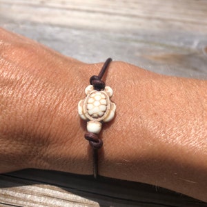 May include: A brown leather bracelet with a white turtle charm. The turtle charm has a detailed shell design.
