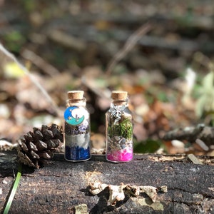 May include: Two small glass bottles with cork tops, filled with glitter, crystals, and dried herbs. The bottle on the left has a blue and green moon charm, while the bottle on the right has a silver fairy charm. Both bottles are sitting on a brown log.