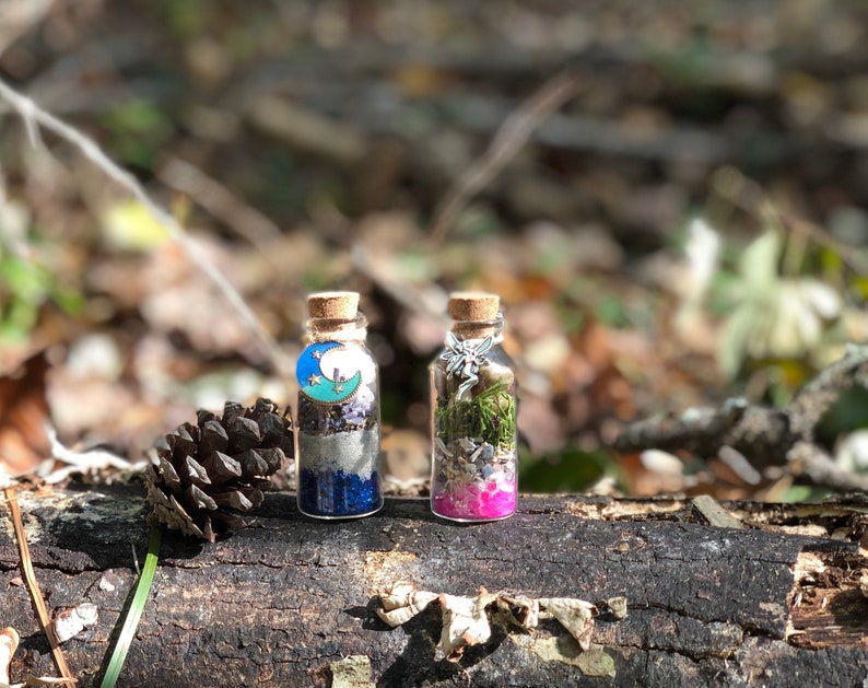 May include: Two small glass bottles with cork tops, filled with glitter, crystals, and dried herbs. The bottle on the left has a blue and green moon charm, while the bottle on the right has a silver fairy charm. Both bottles are sitting on a brown log.