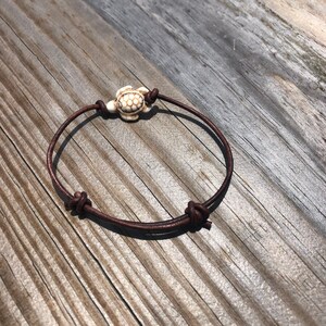 May include: A brown leather bracelet with a white turtle charm. The bracelet is tied with a knot on each end.