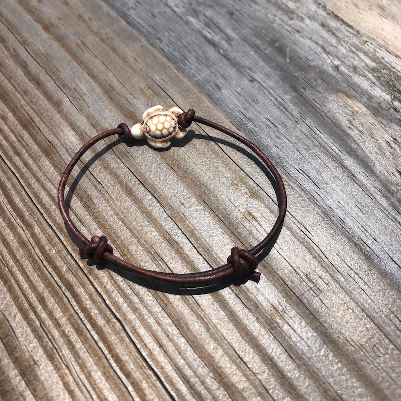 May include: A brown leather bracelet with a white turtle charm. The bracelet is tied with a knot on each end.