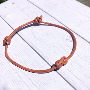 Puede incluir: Una pulsera de cuero marrón claro con tres nudos decorativos. La pulsera está hecha de un cordón de cuero fino y redondo. Los nudos están espaciados uniformemente alrededor de la pulsera. La pulsera está sobre una superficie de madera blanca.