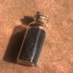 Puede incluir: Una peque&ntilde;a botella de vidrio con un tap&oacute;n de corcho llena de arena negra.