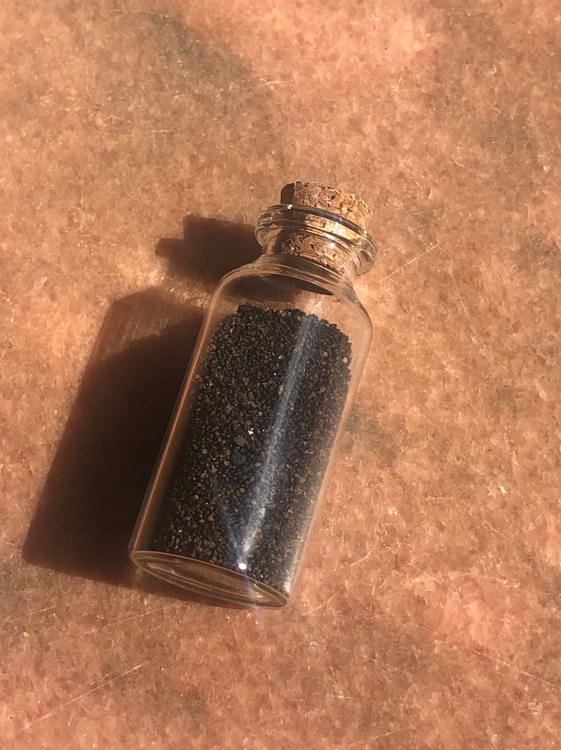Puede incluir: Una peque&ntilde;a botella de vidrio con un tap&oacute;n de corcho llena de arena negra.