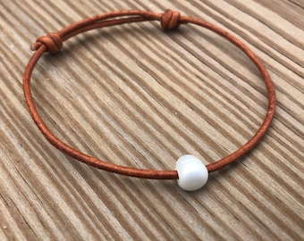 May include: A minimalist bracelet featuring a brown leather cord and a single, white, oval-shaped bead. The cord is knotted at both ends for adjustability. The bracelet is displayed on a wooden surface.