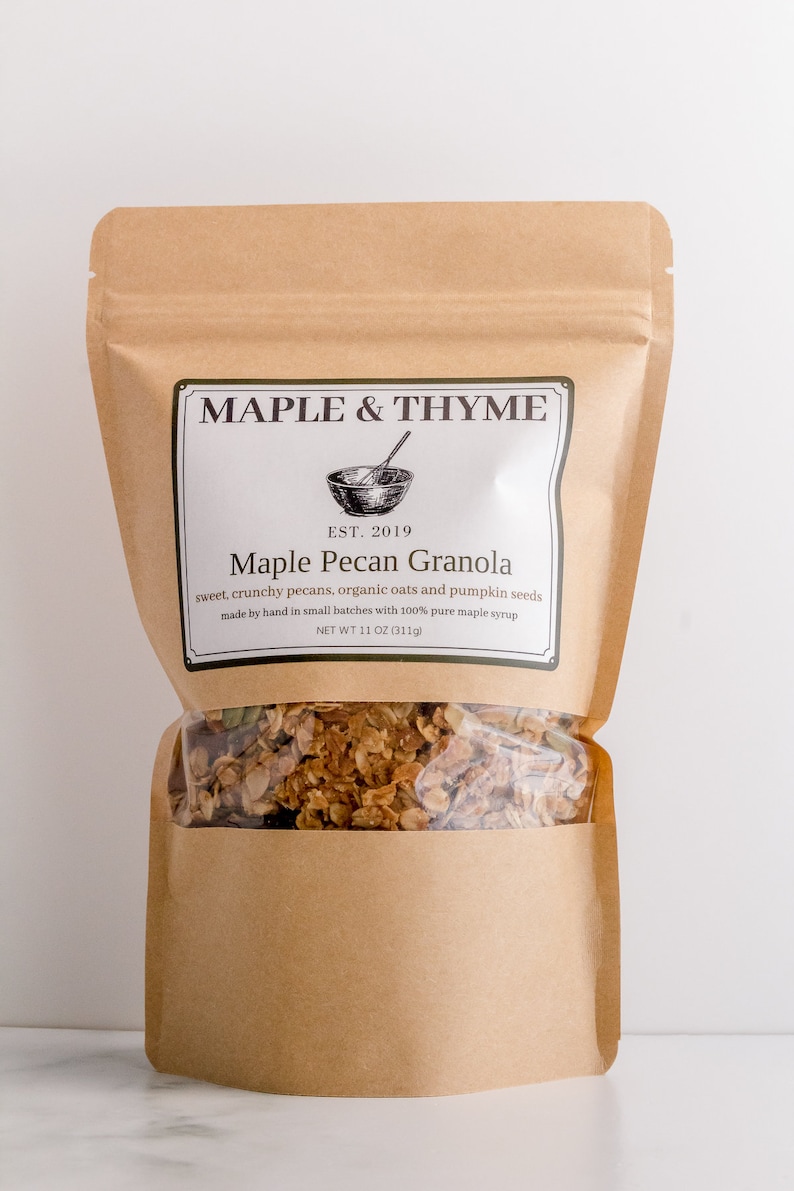 May include: A brown paper bag of Maple Pecan Granola. The bag has a clear window showing the granola inside. The label reads "MAPLE & THYME" and "Maple Pecan Granola". The net weight is 11 oz (311g).