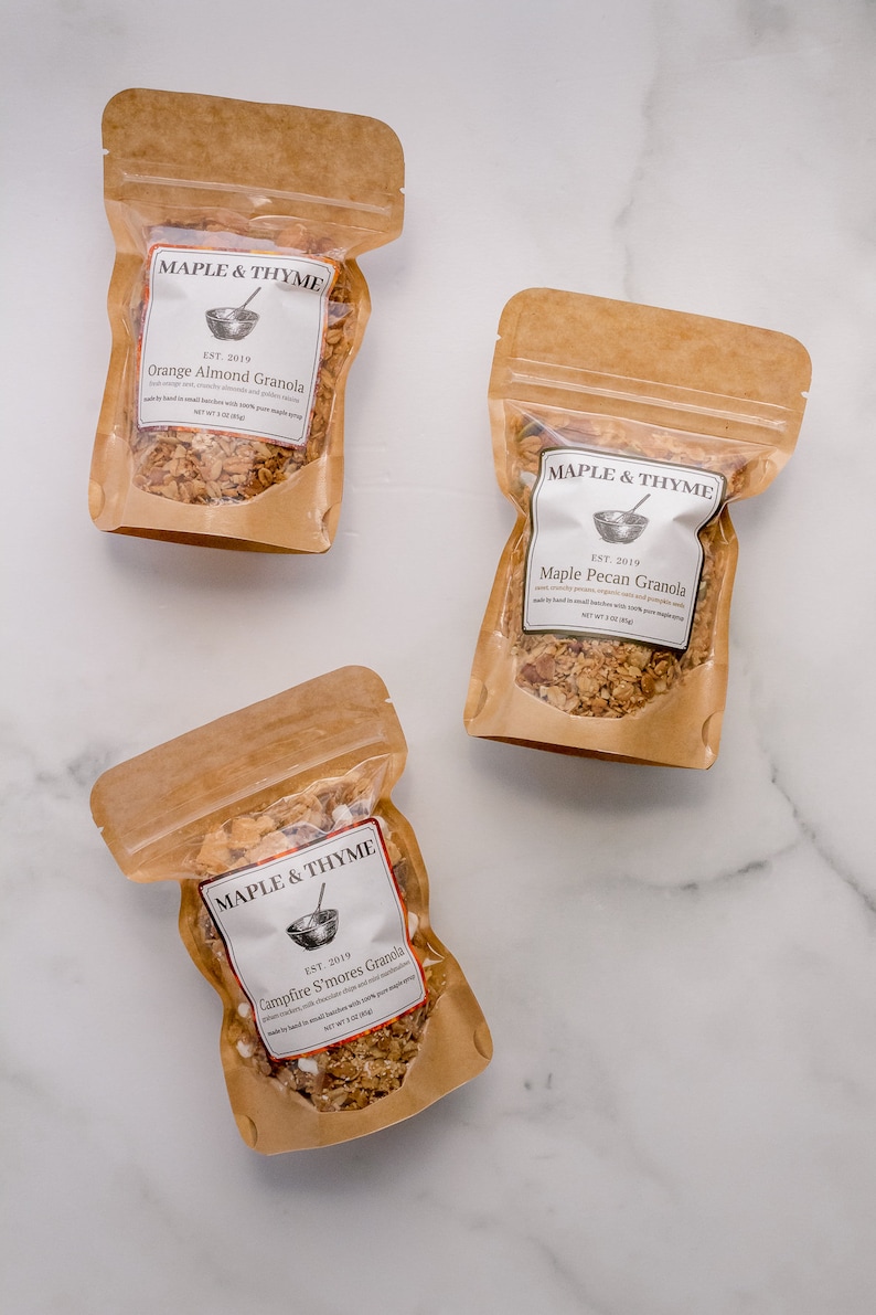 May include: Three brown paper bags with white labels containing different flavors of granola. The labels read "Maple & Thyme" and list the flavors "Orange Almond Granola", "Maple Pecan Granola", and "Campfire S'mores Granola".