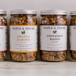 Könnte beinhalten: Vier Glasbehälter gefüllt mit verschiedenen Arten von Granola. Die Behälter sind mit dem Markennamen "Maple & Thyme" und den Geschmacksrichtungen "Campfire S'mores", "Orange Almond", "Cinnamon Raisin" und "Maple Pecan" gekennzeichnet.