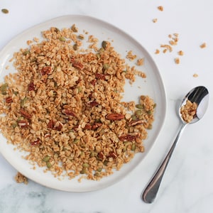 May include: A plate of homemade granola with pecans and pumpkin seeds. The granola is a mix of golden brown oats and nuts, served on a white ceramic plate. A silver spoon rests on the side, filled with granola.