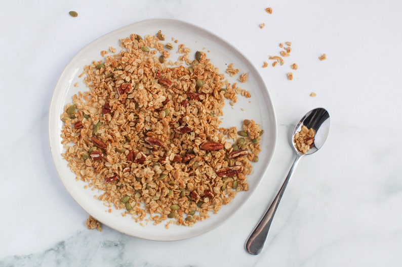 May include: A plate of homemade granola with pecans and pumpkin seeds. The granola is a mix of golden brown oats and nuts, served on a white ceramic plate. A silver spoon rests on the side, filled with granola.