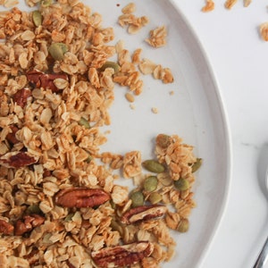 May include: Overhead shot of a white plate filled with granola, featuring oats, pecans, and pumpkin seeds. A silver spoon holds a portion of the granola. The granola is a mix of golden brown and tan colors, with a few pieces scattered on the white surface.