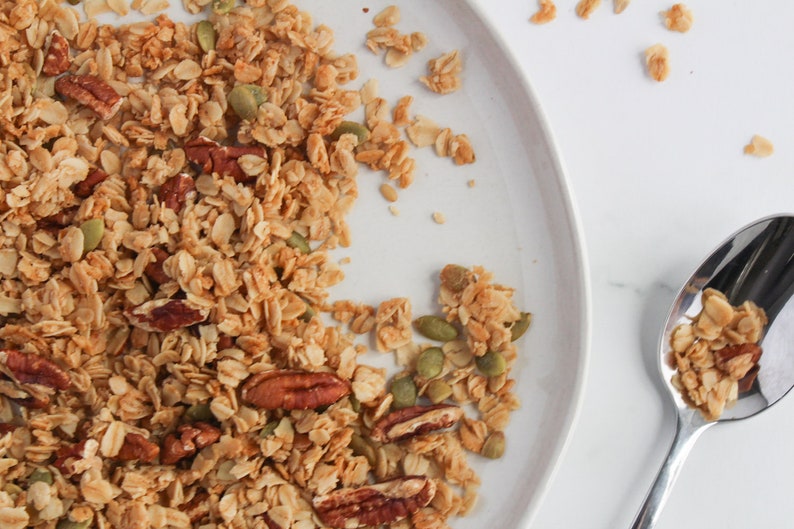 May include: Overhead shot of a white plate filled with granola, featuring oats, pecans, and pumpkin seeds. A silver spoon holds a portion of the granola. The granola is a mix of golden brown and tan colors, with a few pieces scattered on the white surface.